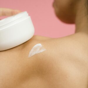Woman applying skincare cream to shoulder against a pink background.
